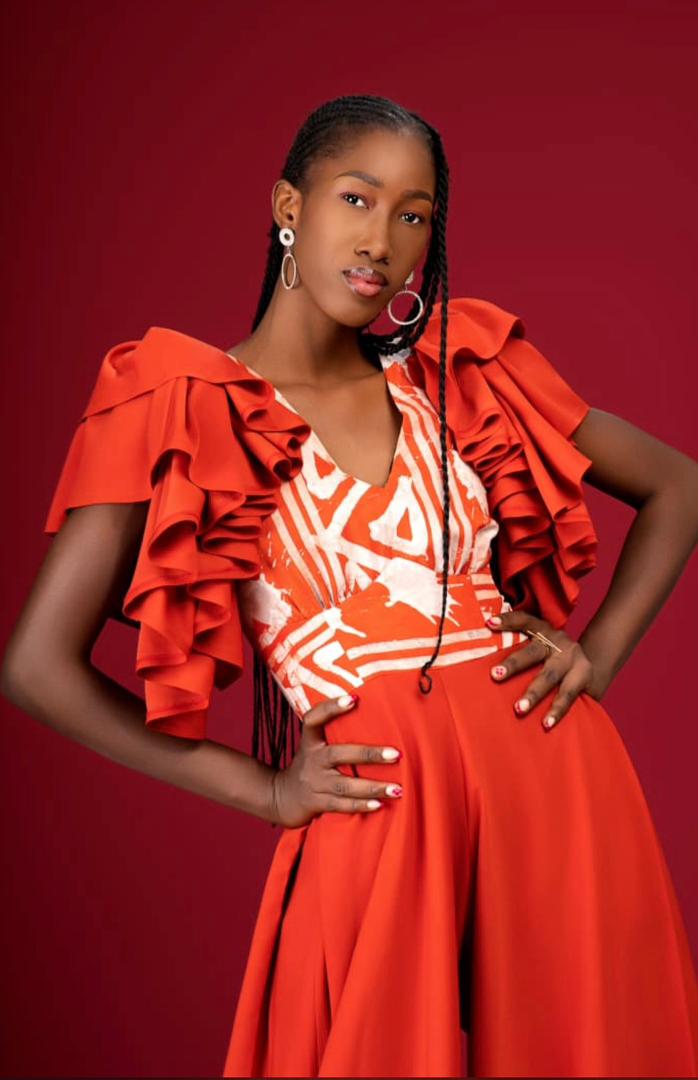 Sakina en robe orange à manches volantées et motifs graphiques blancs, mains sur hanches, fond bordeaux, lumière douce